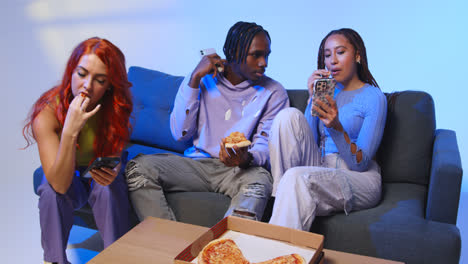 Gen-Z-Friends-Sitting-On-Sofa-Eating-Home-Delivery-Takeaway-Pizza-And-Using-Mobile-Phones